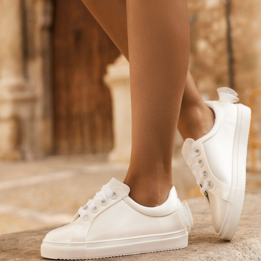 Weißer Brautsneaker mit Schleife, getragen auf einem Steinboden vor einer warmen Hintergrundstruktur. Avalia Shoes bei BAO Warendorf. 