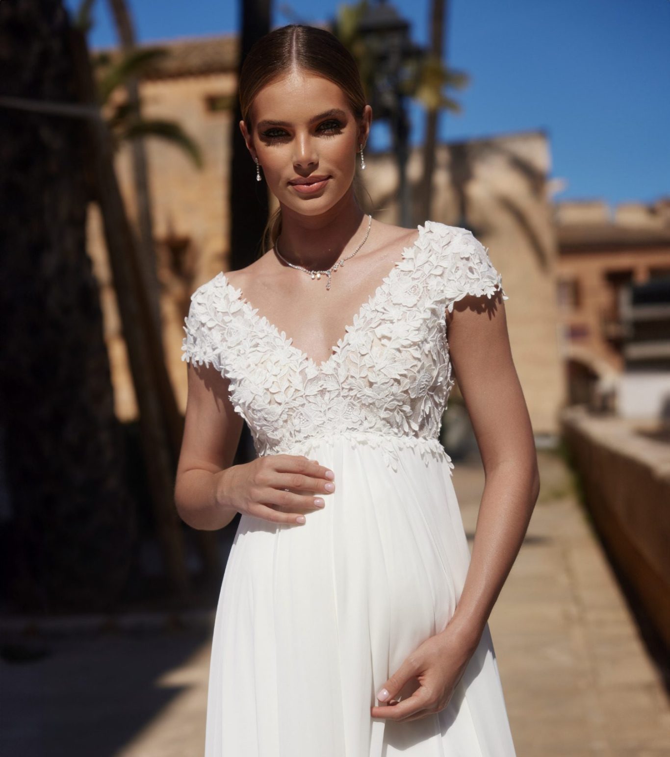 Glückliche Schwangere Braut in einem eleganten weißen Umstandsbrautkleid mit Spitzenverzierung und einer zierlichen Pose. BAO Braut Outlet Warendorf, www.brautoutlet-warendorf.de Schwangere Braut in einem eleganten weißen Umstandsbrautkleid mit Spitzenverzierung und einer zierlichen Pose.